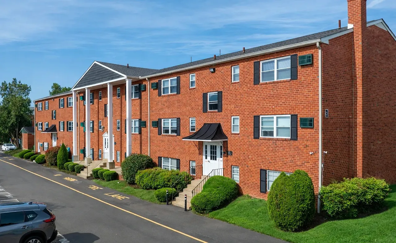A three-story red-brick apartment building with black window shutters and white-trimmed entrances, fronted by a paved drive and landscaped lawn.